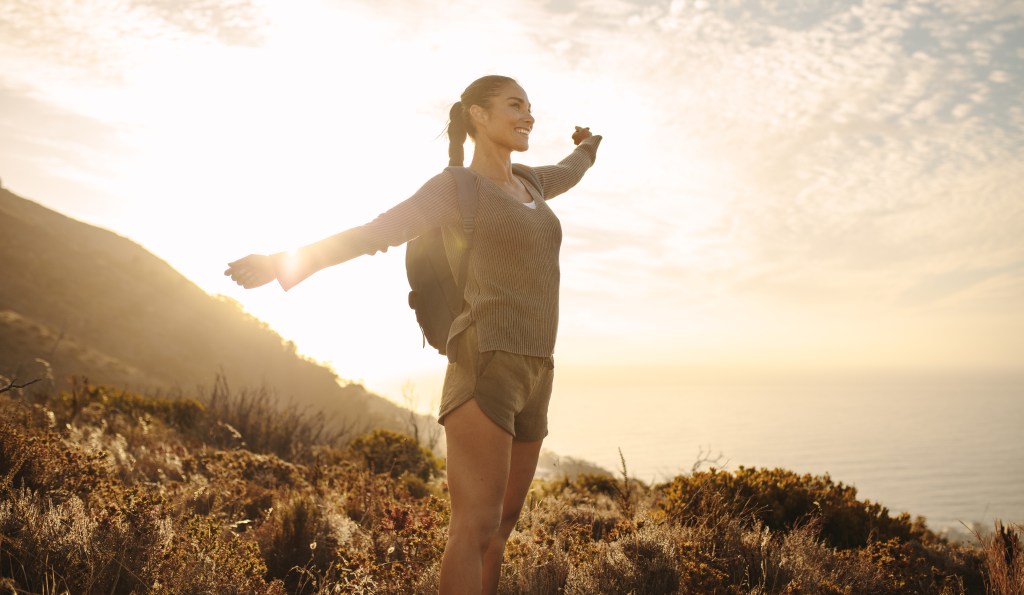 Hiker woman with backpack standing happily on a country path, arms outstretched, in bright sunlight